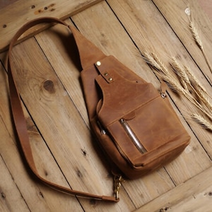 Op de afbeelding: Een bruine leren sling bag met een schouderriem, een zak met ritssluiting en een kleinere voorzak. De tas is gemaakt van distressed leer en heeft messingkleurige hardware. De tas ligt op een houten oppervlak.