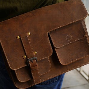 Op de afbeelding: Bruine leren messenger bag met een klepsluiting en meerdere vakken. De tas heeft een schouderriem, metalen hardware en witte stiksels. Het leer heeft een distressed uitstraling.