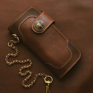 Op de afbeelding: Een bruine leren portemonnee met een decoratieve leeuwenkopsluiting en een goudkleurige ketting. De portemonnee is rechthoekig met een gebogen rand en is gemaakt van getextureerd leer. De ketting is met een sleutelring aan de portemonnee bevestigd.