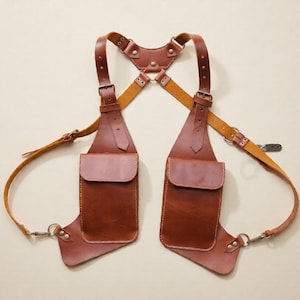 May include: A brown leather shoulder holster with two pouches and adjustable straps. The holster features a cross-body design with metal hardware and a connecting strap at the back. The leather has a rich, warm tone.
