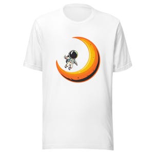 May include: White t-shirt featuring a cartoon astronaut floating on a crescent moon. The astronaut is wearing a white spacesuit and helmet. The moon is orange and yellow. The t-shirt is made of a soft material.