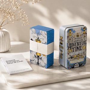 May include: A deck of tarot cards wrapped in a white paper band, a rectangular metal storage tin labelled Totally Unofficial Adventure Time Tarot Deck by Katherine Hillier, and a small white instruction booklet with black text.