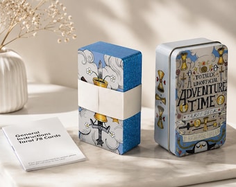 Op de afbeelding: Een set tarotkaarten omwikkeld met een witte papieren band, een rechthoekig metalen opbergblik met het label Totally Unofficial Adventure Time Tarot Deck door Katherine Hillier en een klein wit instructieboekje met zwarte tekst.