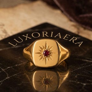 May include: A gold signet ring featuring a square face with rounded corners, engraved with a starburst pattern centred by a small, round, deep red gemstone. The text LUXORIAERA and EXCEPTIONAL ARTISTRY are visible above and below the ring.