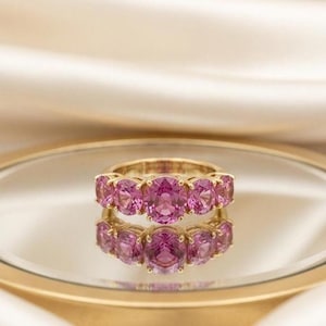 Op de afbeelding: Een gouden ring met vijf ovale, roze edelstenen. De ring wordt gepresenteerd op een reflecterend oppervlak, met de merknaam "Luxoriaera". De achtergrond is een zachte, crèmekleurige stof.