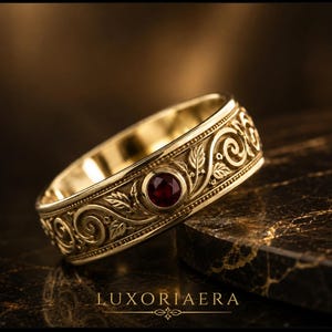 Könnte beinhalten: Ein goldener Bandring mit einem komplizierten Reliefmuster aus geschwungenen Ranken und Blättern. Ein einzelner runder, tiefroter Edelstein ist mittig in die Metallarbeit eingefasst. Der Text LUXORIAERA ist unten im Bild in Gold gedruckt.