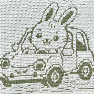 Op de afbeelding: Een wit en saliegroen gehaakt textiel met een grafisch ontwerp van een konijn dat in een kleine auto rijdt. Het konijn zit op de bestuurdersstoel met de oren omhoog, terwijl de auto is afgebeeld met wielen en ramen in het patroon.