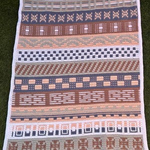 Op de afbeelding: Een gehaakte deken met een geometrisch patroon in wit, perzik, bruin, grijs en blauw. De deken heeft verschillende gestructureerde strepen en een witte rand. Het ontwerp is een mix van herhalende vormen en patronen.