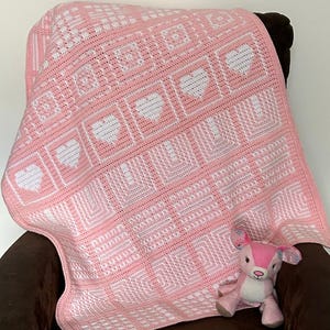 Op de afbeelding: Een rechthoekige gehaakte deken met een herhalend patroon van witte harten en geometrische vierkanten in lichtroze en wit garen. Een klein roze en wit pluche hertje zit op de hoek van de deken die over een stoel gedrapeerd is.