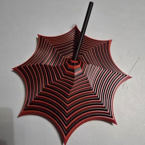 May include: A red and black decorative umbrella-shaped object with a central black pole. The umbrella has a layered design with alternating red and black sections, creating a geometric pattern. The object is likely a decorative piece or a stand.