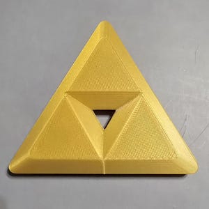 May include: A gold-colored, triangular emblem with a smaller triangle at its center. The emblem has a textured surface and is likely a decorative item or a symbol from a video game.