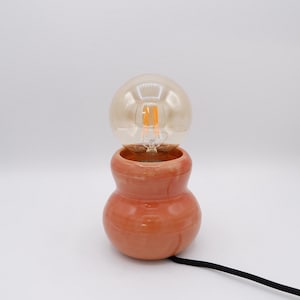 Puede incluir: Una lámpara de mesa con una base de cerámica brillante de color melocotón y forma redondeada de reloj de arena. Una bombilla esférica grande de cristal tintado en ámbar con filamentos internos visibles se asienta en la abertura superior, conectada por un cable negro.