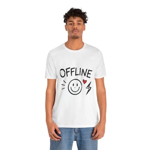 May include: A white short-sleeve t-shirt featuring the black text OFFLINE printed across the chest. Below the text is a black line-drawn smiley face, a small red heart, and a black lightning bolt graphic centered on the front of the garment.