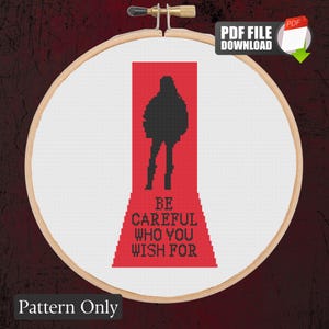 May include: A cross-stitch pattern displayed in a wooden embroidery hoop featuring a black silhouette of a person standing on a red staircase. The text "BE CAREFUL WHO YOU WISH FOR" is stitched in black letters on the red staircase design.