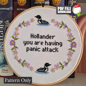 Könnte beinhalten: Ein runder Holzstickrahmen mit weißem Stoff und einem Kreuzstichmotiv. Das Design zeigt einen Blumenkranz aus violetten Blüten und grünen Blättern, zwei schwarz-weiße Eistaucher sowie den Text: Hollander you are having panic attack.