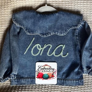 May include: A blue denim jacket with the name "Iona" embroidered in pale green thread. A small square coaster with the text "Embroidery by Mrs. Dowell" and images of yarn and needles is visible.