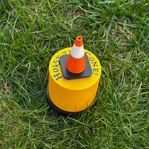 May include: A yellow cylindrical object with the words "HOIST TIE CONE!" printed on the side. A small orange and white traffic cone sits on top of the cylinder. The object is on a bed of green grass.