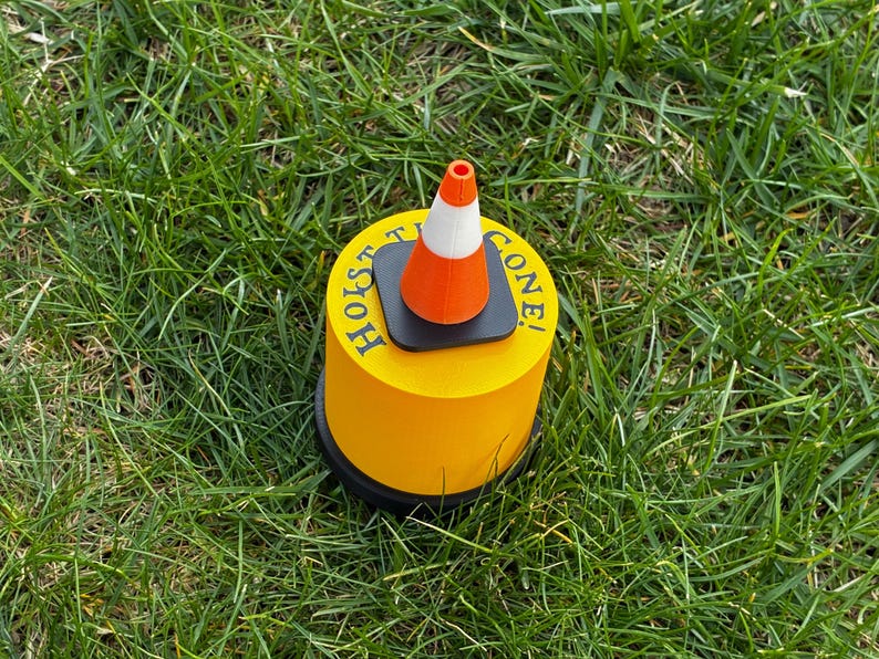 May include: A yellow cylindrical object with the words "HOIST TIE CONE!" printed on the side. A small orange and white traffic cone sits on top of the cylinder. The object is on a bed of green grass.