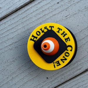 May include: A round, yellow and black pin with the text "HOIST THE CONE!" in black. The pin features an orange and white eye-shaped design in the center.