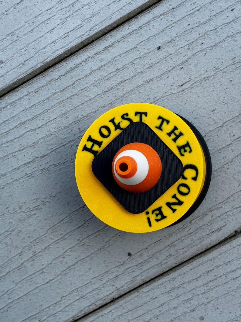 May include: A round, yellow and black pin with the text "HOIST THE CONE!" in black. The pin features an orange and white eye-shaped design in the center.