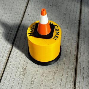 May include: A yellow cylindrical object with the words "Holy Cone!" printed on top, featuring a miniature orange and white traffic cone. The base is black. The cone is a novelty item.