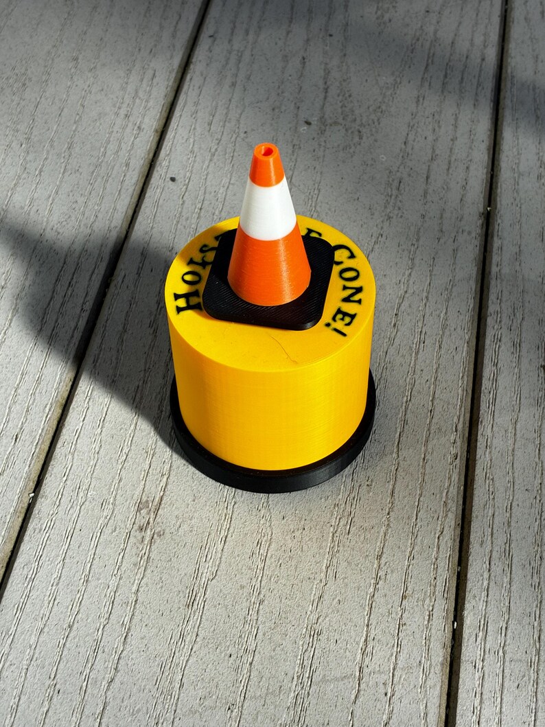 May include: A yellow cylindrical object with the words "Holy Cone!" printed on top, featuring a miniature orange and white traffic cone. The base is black. The cone is a novelty item.