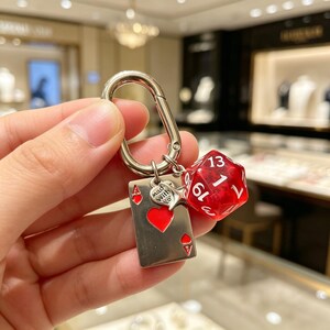 Puede incluir: Llavero plateado con mosquetón, con una carta de juego con un corazón rojo, un dado D20 rojo y un dije que dice "mad with love". La carta de juego es el As de Corazones.