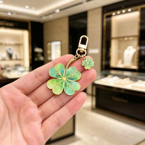 May include: A gold-colored keychain with a four-leaf clover charm and a smaller clover accent. The clover is painted with shades of green and has a gold outline. The keychain is held up by a gold clasp.