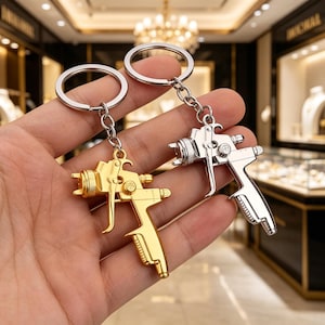 May include: Two keychains with metallic spray gun pendants. One keychain has a gold-colored spray gun, and the other has a silver-colored spray gun. Both keychains have a silver-colored key ring and chain.