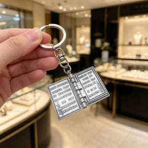 May include: Silver-colored keychain featuring a miniature open book with religious text. The book's pages display engraved script, and the keychain includes a ring and a swivel clasp for attachment. The text on the left page reads "OUR FATHER" and the right page reads "COME THY".