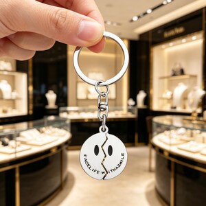Könnte beinhalten: Silberner Schlüsselanhänger mit geteiltem Smiley-Gesicht-Design. Der runde Anhänger trägt die Worte "FACE LIFE WITH A SMILE" am Rand. Das Smiley-Gesicht ist in zwei Hälften geteilt. Der Schlüsselanhänger wird von einer Hand gehalten.