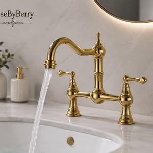 Peut inclure: Un robinet pont doré poli avec deux poignées à levier et un bec à arc élevé, distribuant un jet d'eau constant dans un évier blanc. Le robinet présente un fleuron décoratif sur le dessus et porte l'inscription RoseByBerry.