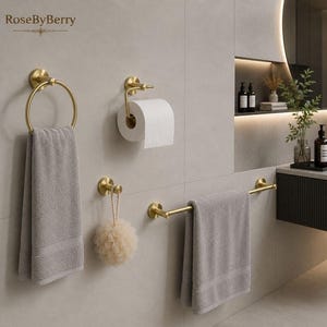 Op de afbeelding: Een set geborsteld goudkleurige badkameraccessoires bestaande uit een ronde handdoekring met een grijze handdoek, een toiletrolhouder, een enkele haak met een beige badspons en een horizontale handdoekstang met een grijze handdoek.