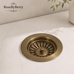 May include: A round kitchen sink drain strainer with a brushed antique brass finish. The metal component features a central knob and a series of radial slots for water drainage. The text RoseByBerry is visible in the upper left corner of the image.