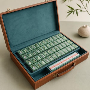 May include: A brown leather-like carrying case with a teal velvet interior containing a set of green mahjong tiles with bamboo and floral designs, alongside a separate row of plain white and pink tiles, featuring a metal handle and latch.