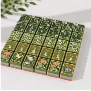 May include: A set of rectangular mahjong tiles arranged in a grid, featuring green surfaces with various floral, bamboo, and numerical designs in white, orange, and black. Each tile has a layered base with a light blue and orange stripe detail.