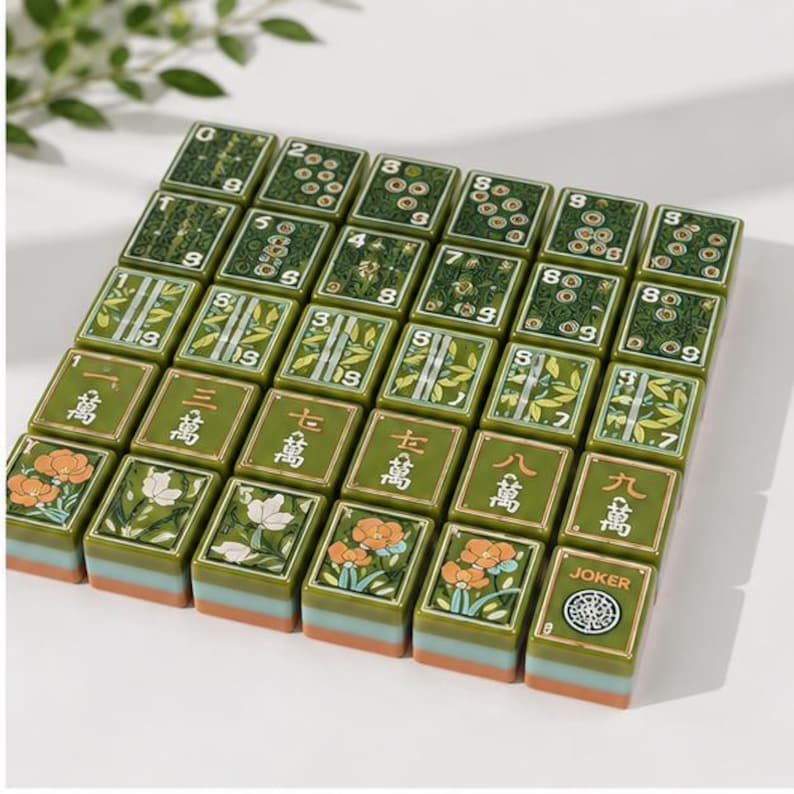 May include: A set of rectangular mahjong tiles arranged in a grid, featuring green surfaces with various floral, bamboo, and numerical designs in white, orange, and black. Each tile has a layered base with a light blue and orange stripe detail.