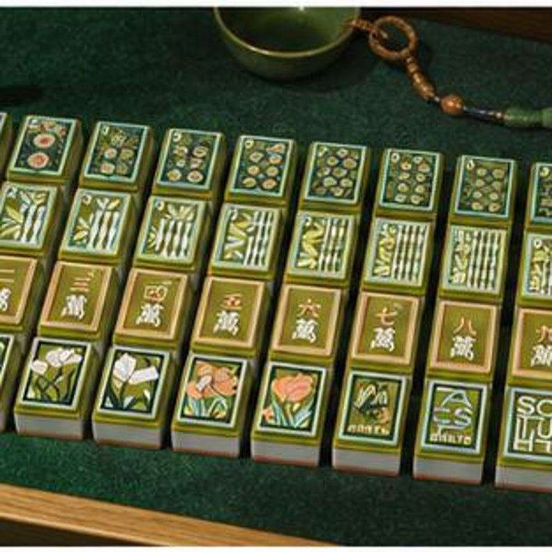May include: A set of rectangular mahjong tiles arranged in rows, featuring green borders and white bases. The tiles display various illustrations including floral patterns, bamboo stalks, and Chinese characters, with some tiles showing the letters A, C, S, and T.