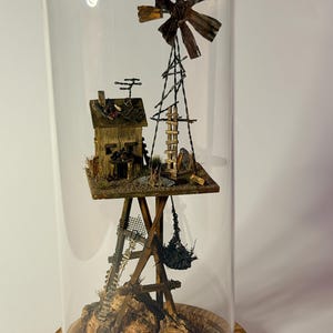 Puede incluir: Un diorama en miniatura de un molino de viento rústico y una pequeña casa, encerrado en una cúpula de cristal. El molino de viento presenta un ventilador de metal marrón desgastado. La casa es de madera y se asienta sobre una plataforma sostenida por una estructura de madera.