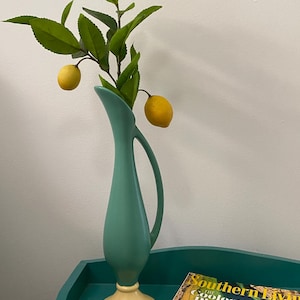 May include: A tall, matte teal vase with a slender neck and a curved handle, featuring a cream-colored circular base. Inside the vase is a green leafy branch with two yellow lemons attached. A magazine with the text Southern Living is visible.