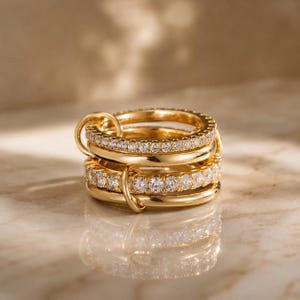 Könnte beinhalten: Ein Stapel aus vier goldenen Ringen, zwei davon mit einem Band aus kleinen, klaren Edelsteinen. Die Ringe sind durch goldene Schlaufen verbunden. Die Ringe werden auf einer reflektierenden Oberfläche mit weicher Beleuchtung präsentiert.