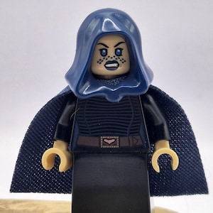 Può includere: Una minifigure Lego di un personaggio con un mantello e un cappuccio blu scuro. La figura ha un abito nero con cintura e un'espressione severa. Il viso ha trucco scuro sugli occhi e piccoli punti sulle guance e sul mento.