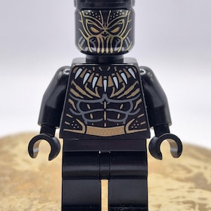 May include: A black plastic toy minifigure featuring a moulded head with pointed ears and gold facial markings. The torso displays a detailed print of a necklace with white teeth and gold chest armour patterns over a black base with grey abdominal lines.