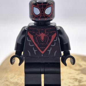 Op de afbeelding: Een zwart plastic minifiguur met een rood spinnenembleem op de borst en een rood webpatroon op het hoofd. Het figuur heeft lichtblauwe ogen, zwarte armen en zwarte benen. Het ontwerp bevat subtiele grijze markeringen op de romp en het hoofd.