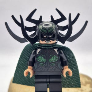 Peut inclure: Une figurine Lego d'un personnage avec un casque noir orné de grandes cornes incurvées. La figurine porte une tenue noire avec des accents verts et une cape verte. La figurine est posée sur une surface brun clair.