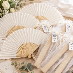 Peut inclure: Plusieurs éventails pliants blanc cassé avec des baleines en bois clair. Certains éventails sont entièrement ouverts, tandis que d'autres sont pliés et emballés dans des sachets en organza blanc. Chaque étiquette porte l'inscription Thank You, To Celebrate With Us.