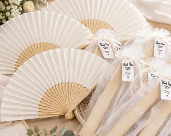 May include: Multiple off-white folding fans with light wood ribs. Some fans are fully opened, while others are folded and enclosed in sheer white organza bags. Each bag features a small white tag with the text Thank You, To Celebrate With Us, and a heart.