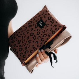 May include: A brown fabric book cover featuring a dark brown leopard print pattern and a black rectangular label with the text Eco Priority. The cover includes a black ribbon tie closure and is shown with a light beige inner lining fabric.