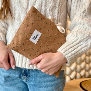 Könnte beinhalten: Ein brauner Stoffbeutel mit einem kleinen Blumenmuster in Gelb und Grün. Auf der Vorderseite ist ein weißes rechteckiges Etikett mit dem schwarzen Text Eco Priority aufgenäht. Der Beutel ist mit einem hellen Schleifenband an der Ecke verschlossen.