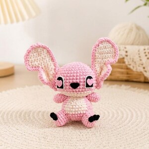 May include: A handmade crochet plush toy in the shape of a pink and white cartoon character with large ears. The toy has black eyes with heart-shaped pupils and black feet. It is sitting on a cream-colored, round rug.